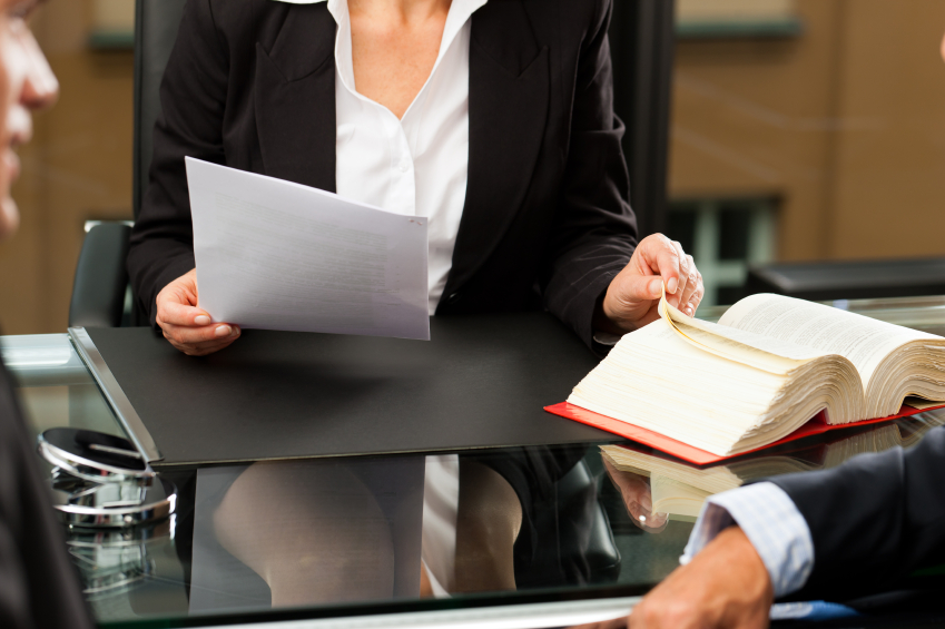 Professionista in tailleur nero alla scrivania di vetro con un foglio in mano sfoglia un grosso codice davanti a due clienti